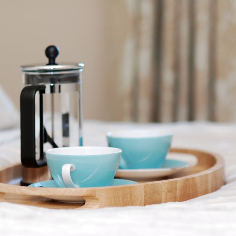 a serving tray with a french press and two cups and saucers on a hotel bed.